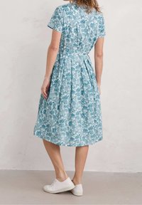 Kobieta ubrana w niebieską i białą, kwiecistą sukienkę do kolan z krótkim rękawem i białymi trampkami, stojąca na tle gładkiej, jasnej ściany.