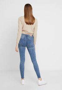 Vaqueros ajustados azules de cintura alta, con dos bolsillos traseros y un parche de cuero marrón en la cinturilla. Combinados con un suéter beige y zapatillas blancas.