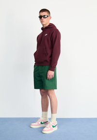 Sweatshirt com capuz na cor vinho, calções verdes e ténis rosa e brancos com detalhes verdes. Inclui um bolso frontal e punhos canelados.