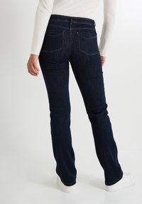 Pantalones vaqueros oscuros de corte slim, con dos bolsillos traseros, costuras sutiles y una cintura alta, combinados con zapatillas blancas.