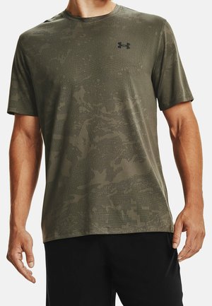 Mężczyzna noszący zieloną, krótką koszulkę sportową Under Armour w kamuflażowy wzór i czarne spodenki, pokazany od szyi do bioder.