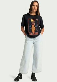Woman standing with hands in pockets wearing black graphic tee with hand and flowers design and "Keep Growing" text, white wide-leg jeans, black boots.