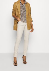 Tan leren dubbelzijdige jas over een blouse met luipaardprint, gecombineerd met lichtbeige getailleerde broek en zwarte haksandalen.