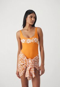 Maillot de bain une pièce orange avec imprimé floral blanc, assorti à un paréo orange et blanc à motifs, noué à la taille.