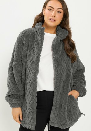 Femme portant une veste polaire grise texturée par-dessus une chemise blanche, la main gauche dans la poche de la veste, sur un fond uni.