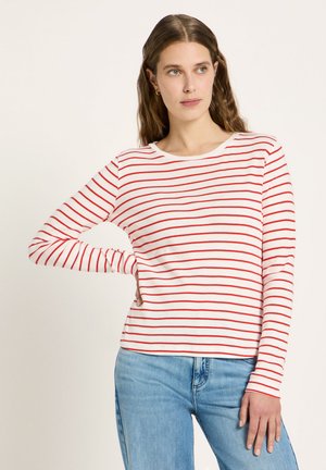 Woman with long wavy hair wearing a white long-sleeve shirt with red horizontal stripes and light blue jeans, standing with one hand on hip.