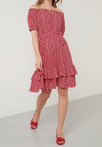 Femme marchant dans une robe rouge à épaules dénudées avec un imprimé floral blanc et un ourlet à volants, associée à des sandales à talons rouges ouvertes à l'avant.