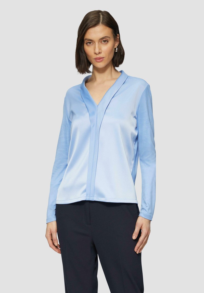 Hellblaues, langärmeliges Blusenshirt mit V-Ausschnitt und vertikalen Nahtdetails. Aus einem glatten Stoff mit lockerer Passform. Kombiniert mit dunklen Hosen.