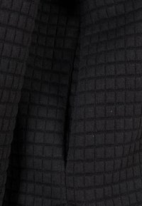 Gros plan sur un tissu noir avec un motif quadrillé texturé et des lignes de couture visibles créant un aspect matelassé.