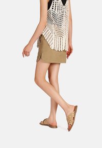 Sandali slide intrecciati marroni con un motivo decorativo, abbinati a una gonna khaki e a un top in crochet crema, con un design libero e aperto.