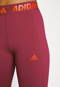 Bordeaux leggings met een brede oranje "ADIDAS" tailleband. Bevat een klein oranje logo op het onderbeen en een gladde, rekbare stof.