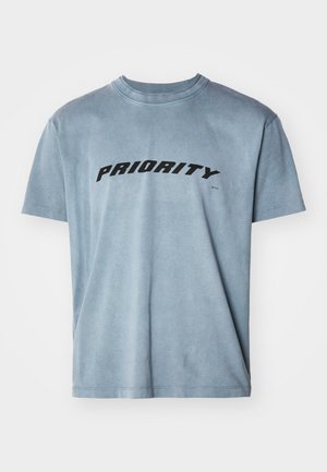 Vaaleansininen puuvillapaita, jossa on lyhyet hihat ja jonka rintamalla on sana "PRIORITY" paksuin mustin kirjaimin. Klassinen pyöreä pääntie.