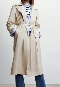 Beiger Trenchcoat mit Taillengürtel, großen Revers und geradem Schnitt. Getragen über einem hellblauen Hemd mit weißen und schwarzen Streifen.