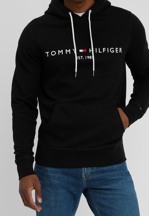 Man draagt een zwarte Tommy Hilfiger hoodie met witte trekkoorden en logo, gecombineerd met een blauwe spijkerbroek, hand in de buidelzak.