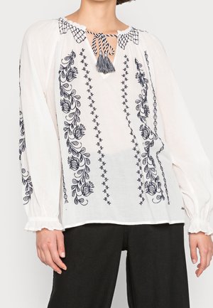 Witte blouse met lange mouwen, met zwarte bloemrijke borduursels, een koord met franjes bij de halslijn en elastische gerimpelde manchetten, gedragen met een zwarte broek.