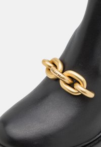Botte en cuir noir avec un large détail chaîne dorée à l'avant, un maillon gravé du logo "COACH".
