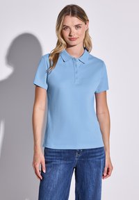 Ljusblå polo med klassisk krage, fyraknappslå, och korta ärmar, i kombination med blå denimjeans.