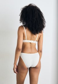 Balta apatinė lingerie su minimalizuotu dizainu, plonomis reguliuojamomis petnešomis, užsegama kabliuko ir akies uždarymu gale ir be siūlių apačia.