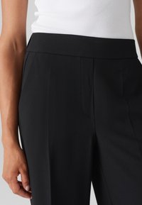 Pantalons noirs ajustés avec une taille plate, des plis latéraux et une texture lisse, montrés de la taille vers le bas avec une main reposant dessus.