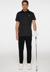 Mustat poolopaita, jossa on kaulus ja lyhyet hihat, yhdistettynä mustiin housuihin ja valkoisiin kenkiin. Golfmaila kädessä oikealla puolella.