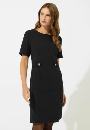 Femme portant une robe noire mi-longue avec des manches courtes et des boutons décoratifs à la taille, debout devant un fond clair uni.