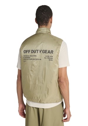 Mand iført beige ærmeløs vest med "OFF DUTY GEAR" og tekst på ryggen, over lyse skjorte med korte ærmer og matchende bukser.