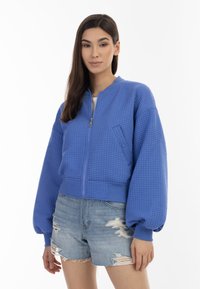 Giacca bomber blu con tessuto testurizzato, chiusura zip frontale, polsini a costine e design cropped. Indossata con pantaloni shorts in denim strappati.