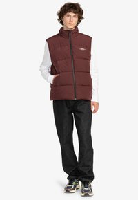 Burgundy pufferväst med hög krage, dragkedjestängning och sidofickor, bärs över en vit långärmad skjorta och kombineras med mörka jeans.