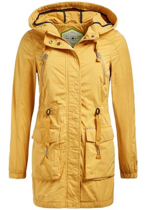 Femme portant une veste jaune à capuche, un jean skinny noir déchiré et des bottines marron, debout les mains dans les poches de la veste.