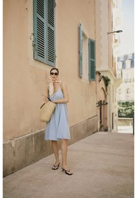 Jasnoniebieska sukienka bez rękawów, pleciona torba ze słomy, czarne okulary przeciwsłoneczne i lekko wzorzysty szalik. W tle beżowa ściana i zielone okiennice.