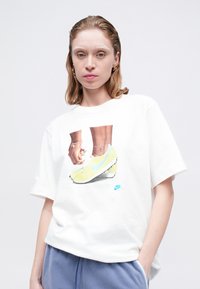 Balts kokvilnas T-krekls ar krāsainu grafiku, kurā attēlotas kedas un rokas. Zils Nike swoosh logo. Brīvs piegriezums ar īsām piedurknēm.