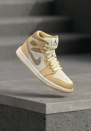 Μπεζ και λευκό αθλητικό παπούτσι Nike Air Jordan μεσαίου ύψους, με κορδόνια, διάτρητο μπροστινό μέρος και λογότυπο φτερών, να αιωρείται πάνω από επιφάνεια από πέτρα.