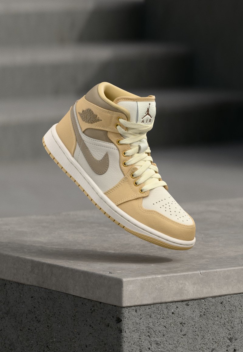 Beige og hvit Nike Air Jordan midt-høy sneaker med lisser, perforert tå og wings-logo, svevende over en steinoverflate.
