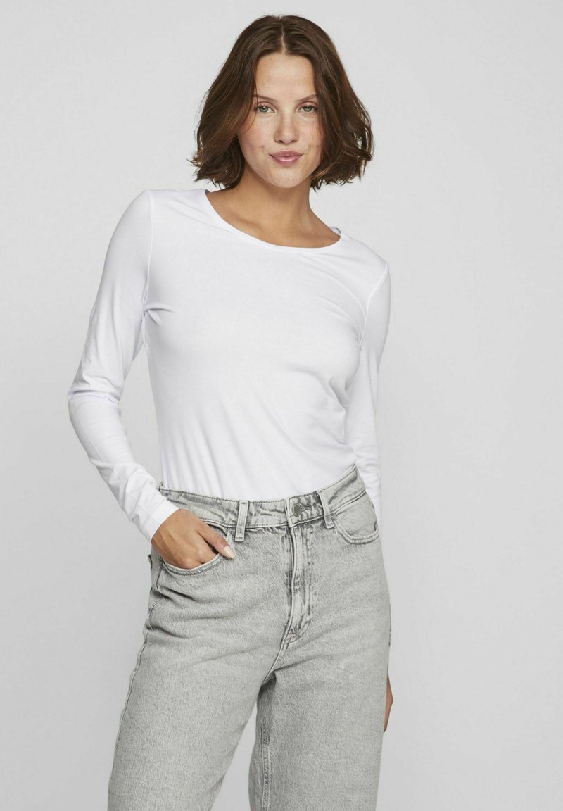Vit långärmad topp med rund halsringning, kombinerad med ljusgrå högmidjade jeans. Slät textur, tajt vid axlarna, avslappnad passform nedanför.