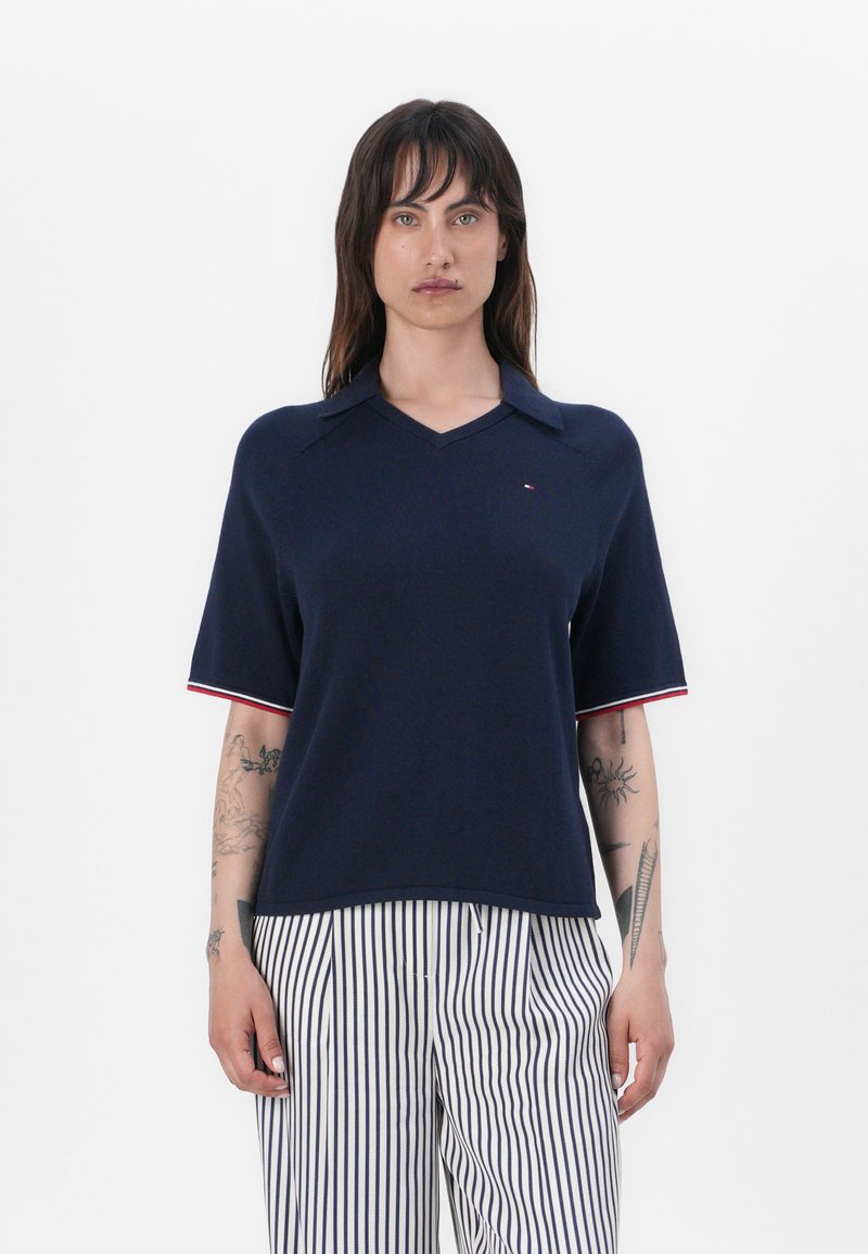 Polo de manga corta en color azul marino con cuello en V, que presenta acentos a rayas blancas y rojas en las mangas y un pequeño logo en el pecho.