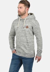 Gray, textured hoodie with a kangaroo pocket, adjustable drawstring, and a small brown logo patch on the chest. Worn with blue jeans.