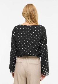 Mustade täpitud pluus pikkade varrukate, tekstureeritud kangaga ja laia kaelusega, mida on kombineeritud beežide kõrge vöökohaga pükstega, millel on tagataskud.