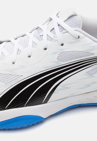 Scarpa sportiva bianca con tomaia in mesh traspirante, caratterizzata da un design con accenti neri e blu. La suola è principalmente bianca con un outsole blu.