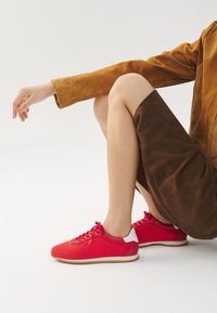 Sneakers rossi con parte superiore in mesh, dettagli bianchi sul tallone e lacci. Posizionati su uno sfondo neutro, abbinati a una gonna in suede marrone.