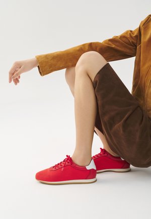 Red sneakers with a mesh upper, white heel accents, and laces. Set against a neutral background, paired with a brown suede skirt.