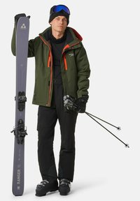 Skiausrüstung mit einer grünen Jacke mit orangefarbenen Akzenten, schwarzen Skihosen, schwarzen Handschuhen und grauen Skiern mit Bindungen. Schwarze Stiefel und Skistöcke sind ebenfalls enthalten.