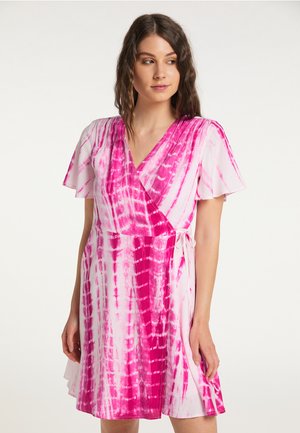 Vrouw draagt een korte mouw, roze en witte tie-dye wikkeljurk met V-hals, kijkt naar de zijkant tegen een effen lichte achtergrond.