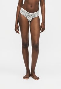 Γυναίκα που στέκεται ξυπόλητη φορώντας λευκά δαντελωτά μαγιό-μπικίνι μπροστά σε απλό φωτεινό φόντο.