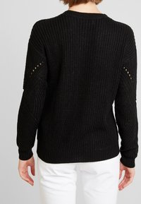 Eine Person trägt einen strukturierten schwarzen Pullover mit offenen Strickdetails an den Ärmeln, kombiniert mit weißen Hosen und blickt von der Kamera weg.
