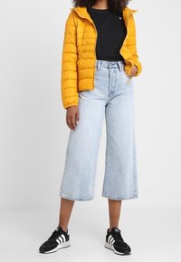 Żółta kurtka puchowa, czarna koszulka, jasnoniebieskie dżinsy o szerokich nogawkach i czarne sneakersy. Gładka tekstura i widoczne szwy na kurtce.