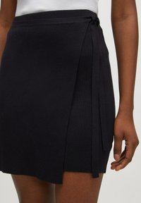 Gonna nera a coste con dettaglio annodato lateralmente. Presenta una linea dritta e un tessuto dalla texture evidente. Mano visibile che tiene il tessuto sul fondo.