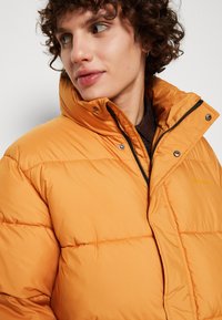 Oranje pufferjack met een hoge kraag, ritssluiting en drukknoopsluitingen. Heeft een zachte, gewatteerde textuur en subtiele branding.