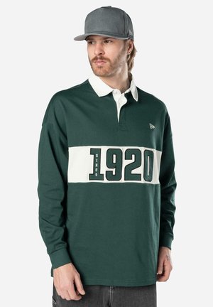 Mann mit hellbraunen Haaren und Bart trägt eine graue Kappe und ein dunkelgrünes Rugbyshirt mit weißem Kragen und der Aufschrift „SINCE 1920“ auf der Brust.