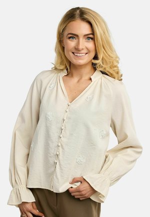 Usmievajúca sa žena s blond vlasmi nosí krémovú blúzku s kvetinovou výšivkou, detailmi na gombíkoch a volánikovými manžetami, pózujúc na bielom pozadí.