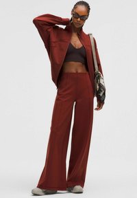 Bordó tréningruha szett, amely zipzáras dzsekiből és széles szárú nadrágból áll, barna sportmelltartóval és világosszürke sportcipővel párosítva.